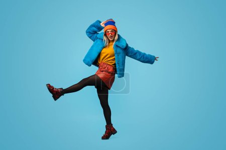 Foto de Longitud completa extraña joven en traje elegante colorido levantando la pierna y tocando el sombrero mientras baila sobre fondo azul - Imagen libre de derechos