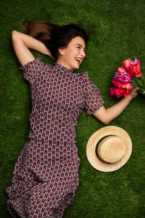 Foto de Desde arriba mujer joven en vestido elegante y sombrero que sostiene ramo de tulipanes mientras está acostado en la hierba verde - Imagen libre de derechos