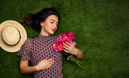 Téléchargez les photos : Sensuelle jeune femme en tenue décontractée tenant tas de tulipes tout en étant couché herbe verte près du chapeau en été avec espace de copie - en image libre de droit