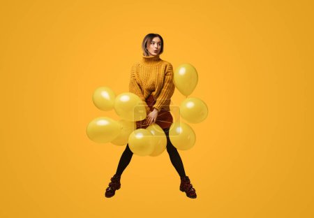Foto de Sonriente joven hembra en traje de moda saltando con un montón de globos brillantes sobre fondo amarillo - Imagen libre de derechos