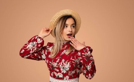 Photo for Astonished young chic in stylish floral dress covering mouth with hand and looking away while standing against beige background - Royalty Free Image