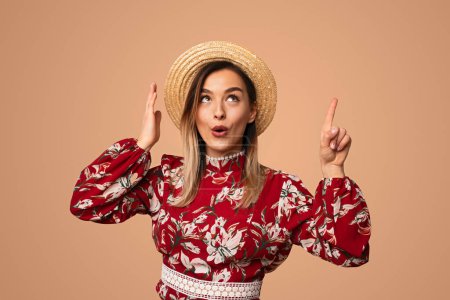 Téléchargez les photos : Jeune femme étonnante en robe florale tendance et chapeau de paille pointant vers le haut tout en se tenant debout sur fond beige - en image libre de droit