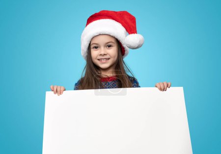 Foto de Chica feliz en el sombrero de Santa sosteniendo la bandera blanca en blanco y mirando a la cámara mientras sonríe contra el fondo azul - Imagen libre de derechos