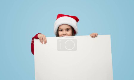 Téléchargez les photos : Joyeux petite fille dans Santa chapeau souriant et regardant la caméra tout en démontrant bannière vierge sur fond bleu - en image libre de droit