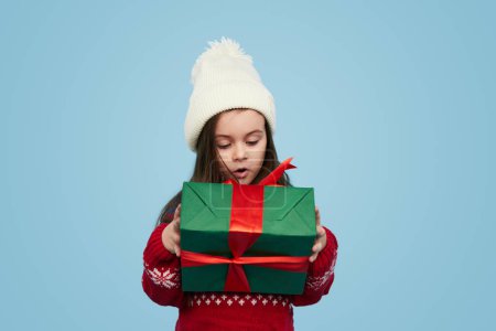 Foto de Muchacha de niño sorprendido en ropa de abrigo y sombrero unboxing una caja de regalo de colores sobre fondo azul - Imagen libre de derechos