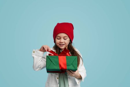 Foto de Feliz niña preadolescente en traje de moda y caja de regalo de apertura de sombrero rojo mientras sonríe contra el fondo azul - Imagen libre de derechos