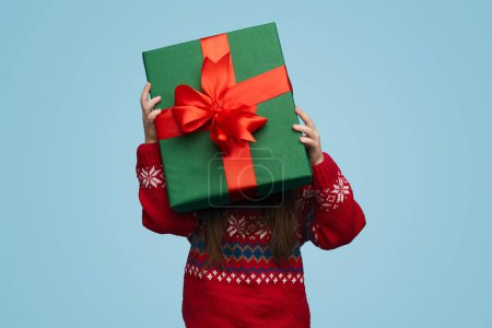 Foto de Niña irreconocible en jersey rojo sosteniendo una gran caja de regalo y cubriendo su cara contra fondo azul - Imagen libre de derechos
