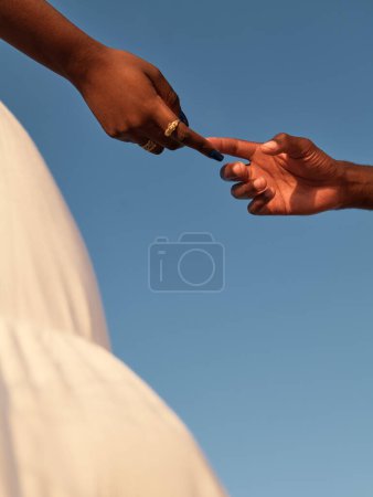 Photo for From below of anonymous ethnic pregnant female in white dress, while standing with faceless male touching index fingers together against cloudless blue sky in sunny daylight - Royalty Free Image