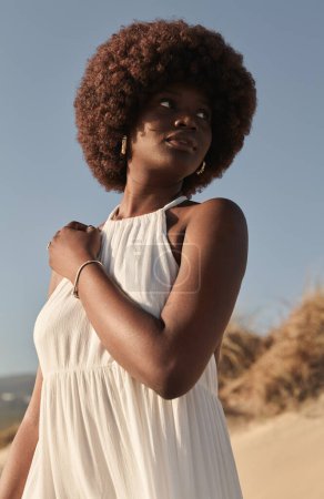 Téléchargez les photos : Angle bas de la jeune femme afro-américaine en robe blanche et avec la coiffure afro détournant les yeux, tout en se tenant debout dans la lumière du jour ensoleillée contre la nature floue et le ciel bleu sans nuages - en image libre de droit