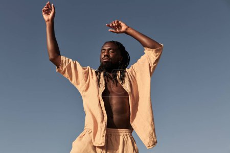 Foto de Bajo ángulo de macho africano con rastas levantando brazos mientras baila contra el cielo azul sin nubes a la luz del sol - Imagen libre de derechos