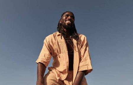 Foto de Bajo ángulo de hombre negro joven feliz en traje beige de pie contra el cielo azul sin nubes en el día soleado y riendo - Imagen libre de derechos
