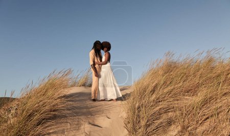 Téléchargez les photos : Vue latérale du jeune couple afro-américain en tenues décontractées tout en se tenant étroitement sur le sable avec herbe sèche femelle, regardant caméra contre ciel bleu sans nuages par jour ensoleillé - en image libre de droit