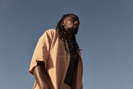 Photo for Low angle side view of confident young African American male in braid hairstyle looking away, while standing alone in daylight under cloudless blue sky in summertime - Royalty Free Image