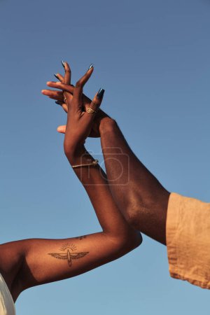 Téléchargez les photos : Couple ethnique anonyme tandis que la femelle debout avec tatouage entrelacant les doigts du mâle et serrant les mains ensemble contre ciel bleu sans nuages en plein jour ensoleillé - en image libre de droit