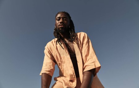 Photo for Low angle of bearded young African American male with braid hairstyle looking at camera, while standing alone with bent posture against cloudless blue sky in sunny daylight - Royalty Free Image