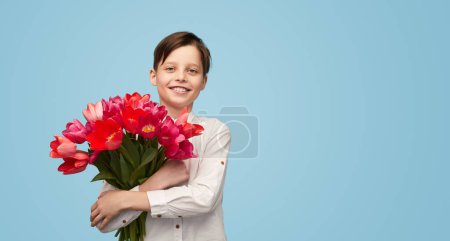 Photo for Happy little boy in white shirt embracing big bunch of blooming spring tulips for 8 March holiday smiling at camera - Royalty Free Image