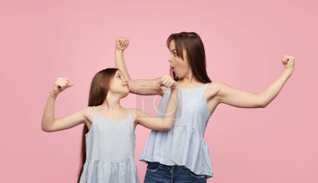 Téléchargez les photos : Heureuse femme adulte avec une petite fille portant des vêtements similaires et montrant biceps posant joyeusement sur fond rose - en image libre de droit