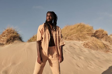 Foto de Joven chico negro con rastas en ropa de verano beige de pie en la costa de arena con hierba seca y mirando hacia otro lado pensativamente - Imagen libre de derechos