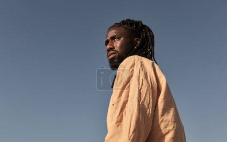 Foto de Bajo ángulo de hombre africano barbudo joven en camisa arrugada de pie contra el cielo azul sin nubes a la luz del sol mirando hacia otro lado - Imagen libre de derechos