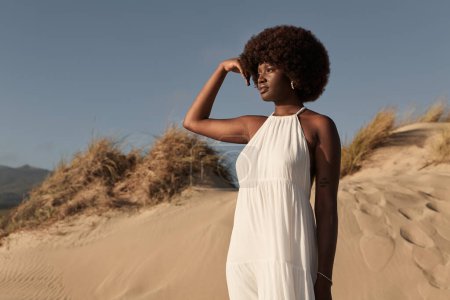 Foto de Joven mujer africana en vestido blanco de verano tocando el cabello afro y mirando hacia otro lado pensativamente mientras está de pie sobre la arena contra el cielo azul sin nubes a la luz del sol - Imagen libre de derechos
