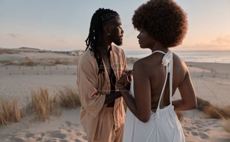 Foto de Joven hombre africano con rastas de pie en la orilla del mar y tomados de la mano con su novia en vestido blanco contra la puesta del sol sobre el mar - Imagen libre de derechos