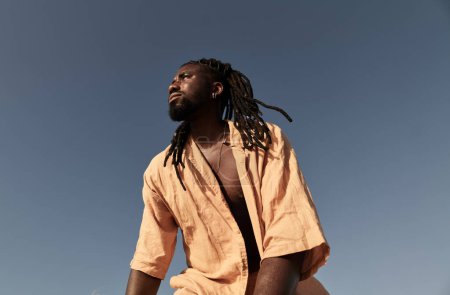 Foto de Bajo ángulo de hombre negro en camisa de verano con rastas de pie contra el cielo azul sin nubes y mirando hacia otro lado en el día soleado - Imagen libre de derechos