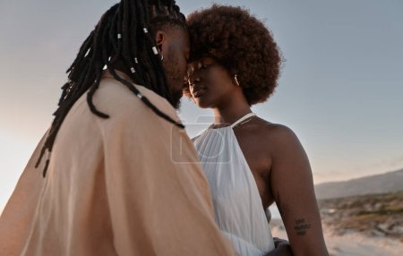 Photo for Low angle of black man with dreadlocks embracing girlfriend in white dress while standing close to each other on seashore against sunset sky - Royalty Free Image