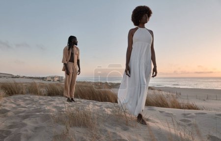 Photo for Full body of young African man standing on sandy hill behind girlfriend in white dress against sea under sunset sky - Royalty Free Image