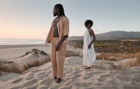 Foto de Cuerpo completo de mujer africana con pelo afro en vestido blanco de pie detrás de novio en la orilla arenosa cerca del mar contra las montañas al atardecer - Imagen libre de derechos
