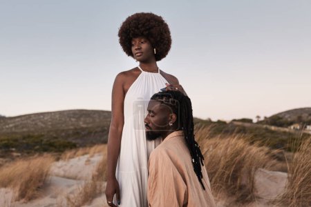 Foto de Joven hombre africano sensual con rastas arrodillado y abrazando novia en vestido blanco de pie en la colina cubierta de hierba seca contra el cielo puesta del sol - Imagen libre de derechos