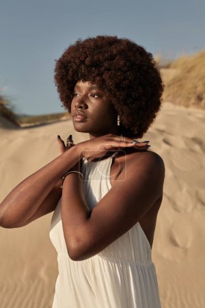 Foto de Joven dama africana reflexiva con pelo afro en vestido blanco cruzando las manos en el pecho mientras está de pie en la playa de arena contra el cielo azul sin nubes - Imagen libre de derechos