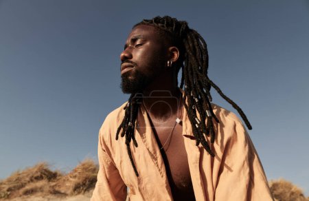 Photo for Low angle of young African man with dreadlocks and closed eyes standing on hill against cloudless blue sky in sunlight - Royalty Free Image