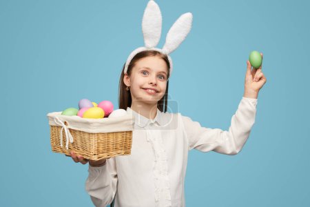 Foto de Encantadora chica de orejas de conejo blanco sosteniendo canasta de mimbre con huevos de colores posando sobre fondo azul - Imagen libre de derechos