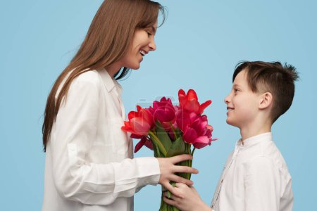 Foto de Vista lateral del niño dando hermoso ramo de tulipanes a la madre de pie juntos sobre fondo azul - Imagen libre de derechos