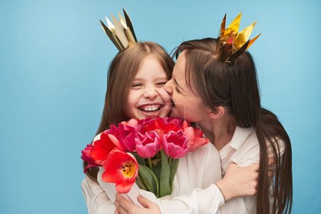 Photo for Woman kissing charming daughter in cheek while both wearing golden crowns and holding 8 March bouquet - Royalty Free Image