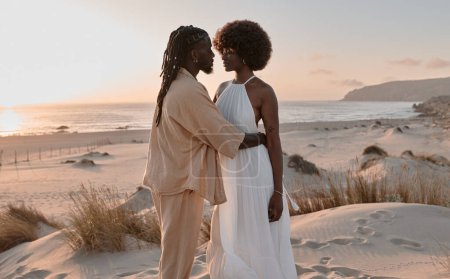 Foto de Joven pareja afroamericana con peinado afro y vestidos de verano, mirándose mientras están de pie en el campo de arena con hierba seca y abrazándose contra el cielo azul nublado y el agua de mar - Imagen libre de derechos