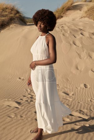 Téléchargez les photos : Vue latérale de la jeune femme afro-américaine avec coiffure afro et robe d'été, tout en se tenant les yeux fermés sur le champ de sable et la main touchant le ventre contre la nature floue et le ciel bleu - en image libre de droit