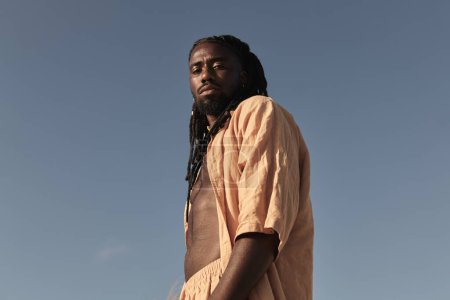 Foto de Bajo ángulo de curioso joven afroamericano masculino con peinado afro trenza y en vestido de verano, mirando a la cámara mientras está de pie solo contra el cielo azul sin nubes en la luz del día soleado - Imagen libre de derechos