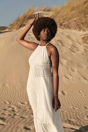 Foto de Vista lateral de la joven afroamericana con peinado afro y vestido de verano, mirando hacia otro lado mientras está de pie en el campo de arena y con la mano tocando el pelo contra la naturaleza borrosa y el cielo azul - Imagen libre de derechos