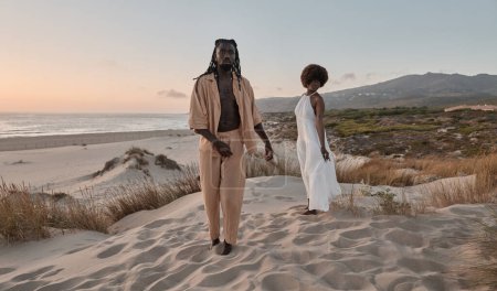 Foto de Joven pareja afroamericana con peinado afro y vestidos de verano, mientras camina descalzo macho mirando a la cámara y de pie hembra en la arena mirando hacia el cielo nublado y el agua de mar - Imagen libre de derechos