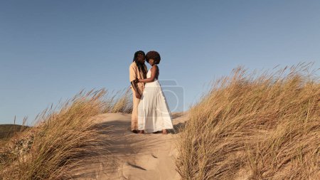 Foto de Joven pareja afroamericana con peinado afro y vestidos de verano, mirando a la cámara mientras está de pie de cerca en el campo de arena con plantas de hierba seca contra el cielo azul sin nubes - Imagen libre de derechos