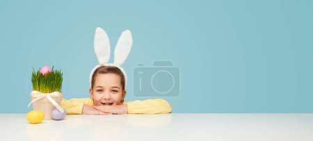 Foto de Adorable niña en orejas blancas apoyada en las manos a la mesa con huevos de Pascua de colores y maceta - Imagen libre de derechos