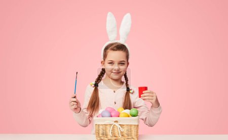 Foto de Niña contenta sentada a la mesa con pinturas y huevos de Pascua para colorear sobre fondo rosa - Imagen libre de derechos