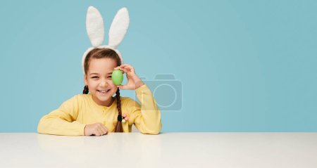 Photo for Expressive child girl wearing white bunny ears and covering eye with colored egg on blue background - Royalty Free Image
