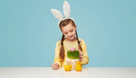 Foto de Chica feliz con trenzas con orejas de conejo blanco y encontrar huevos de Pascua en la hierba de la maceta - Imagen libre de derechos