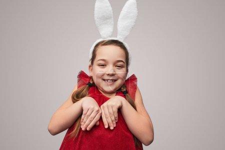 Foto de Encantadora chica en vestido rojo y orejas blancas haciéndose pasar por lindo conejito de Pascua sobre fondo gris - Imagen libre de derechos