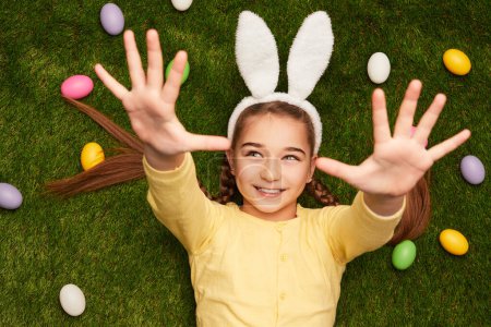Foto de Vista superior de la encantadora chica en orejas de conejo acostado en el césped con huevos de colores extendiendo las manos hacia la cámara - Imagen libre de derechos