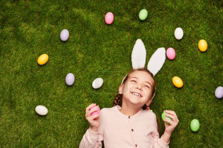 Foto de Desde arriba de niña usando orejas de conejo blanco y acostada sobre hierba verde entre los huevos de Pascua - Imagen libre de derechos