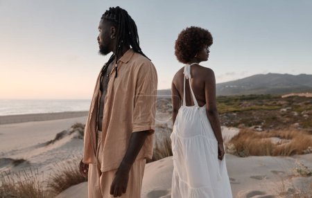 Photo for Side view of averse young African American couple with Afro hairstyle and in summer dresses, looking away in different directions while on sandy beach with back to back against cloudy sky and seawater - Royalty Free Image
