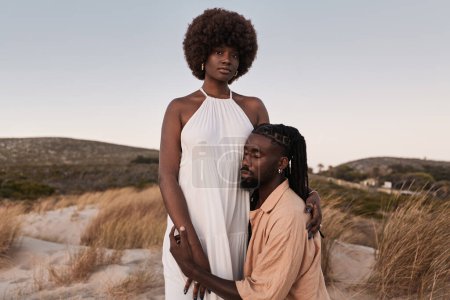 Photo for Side view of young African American male with Afro hairstyle and in summer dress, while sitting with eyes closed with head near belly of standing female looking at camera on grassy sand - Royalty Free Image